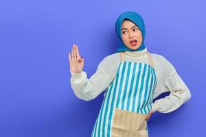 Portrait of angry young Asian Muslim woman in hijab and striped apron showing stop gesture with palm refusing say no isolated on purple background. People housewife muslim lifestyle concept photo