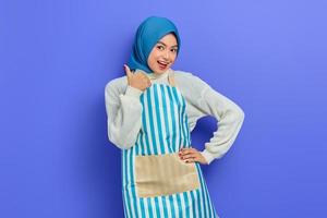 Portrait of cheerful young housewife woman in hijab and striped apron showing thumbs up, looking camera isolated on purple background. People housewife muslim lifestyle concept photo