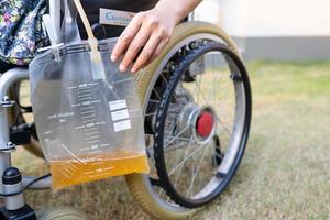 Asian lady patient sitting on wheelchair with urine catheter drainage bag in hospital ward, healthy medical concept. photo