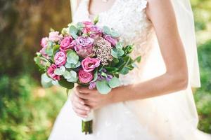 bride in a white dress holding a bouquet of purple flowers and greenery on the background of green grass photo