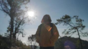 una turista femminile che indossa una giacca gialla guarda i rami di un grande albero su uno sfondo di cielo blu in una foresta tropicale autunnale. giovane donna felice ed eccitata nell'avventura all'aria aperta video