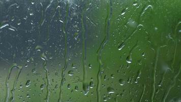 Background with macro shot of a glass of a house window with falling rain drops during heavy summer rain with blurred green trees outside. 4K resolution video.