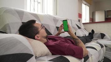 Junger Mann liegt auf der Couch und starrt auf sein Handy, grüner Bildschirm