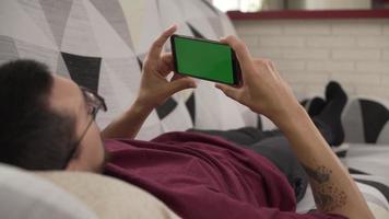 jeune homme allongé sur le canapé regardant fixement son téléphone portable, écran vert video