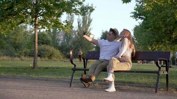 Young couple taking a selfie on their mobile sitting in the park. Latin man and Caucasian woman video