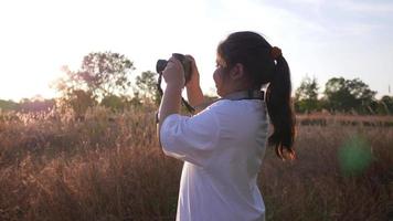 heureuse fille asiatique étant dans le pré avec appareil photo, prenant une photo de vue. debout sur l'herbe en belle journée avec fond de ciel bleu. lumière du soleil merveilleuse video