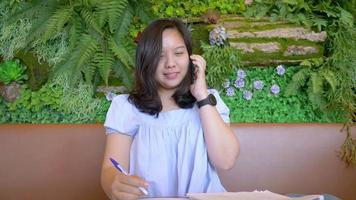 asiatische Frauen, die in einem Café sitzen und arbeiten und ein Telefon abheben, Geschäftsfrau, die versucht, Startup-Geschäftsideen und -pläne im Café zu schreiben, kreative Gedanken in ein Notizbuch. Listenkonzept erstellen video