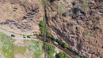 Aerial drone view of people bathing on a high waterfall falling on the road on a sunny day. Cars passing. Anjos waterfall in Madeira Island.  Hot day people shower below the waterfall. Travel.