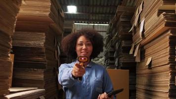 una lavoratrice afroamericana in uniforme di sicurezza che utilizza uno scanner di codici a barre davanti a una telecamera, controlla gli ordini di spedizione nel magazzino dei pacchi, una fabbrica di produzione di cartone per l'industria dell'imballaggio.