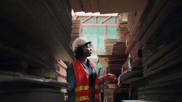 An African American Female worker in safety uniform and hard hat, supervisor inspector packaging stock order at factory warehouse, piles of stacking paper manufacture, recycling production industry.