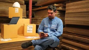 Asian male worker in safety uniform sit and mobile phone chatting to check shipment orders at parcels warehouse, cardboard manufacture factory for the packing industry, logistic transport service. video