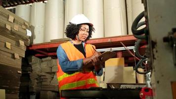 una trabajadora industrial afroamericana con un sombrero duro revisa la máquina y escribe las piezas del motor inspeccionadas. trabajos profesionales en una fábrica de fabricación de papel, mantenimiento de equipos de producción. video