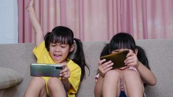 dos hermanas están jugando juegos en línea en sus teléfonos inteligentes sentadas en el sofá de casa. concepto moderno de comunicación y adicción a los gadgets. dos niños con aparatos. video