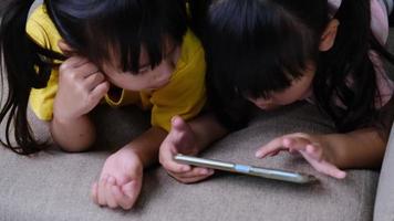 dos hermanas están jugando juegos en línea en teléfonos inteligentes acostados en el sofá en casa. concepto moderno de comunicación y adicción a los gadgets.