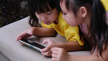 deux sœurs jouent à des jeux en ligne sur leurs smartphones assis sur le canapé à la maison. concept moderne de communication et de dépendance aux gadgets. deux enfants avec des gadgets. video