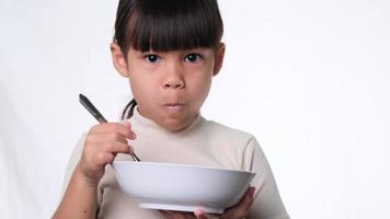 söt liten flicka som äter frukost. glad liten flicka äter spannmål med mjölk från skål på vit bakgrund i studio. hälsosam kost för barn. video