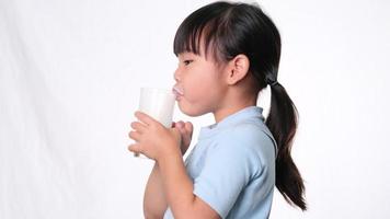 linda niña asiática bebiendo leche de un vaso y lamiendo sus labios sobre fondo blanco en el estudio. nutrición saludable para niños pequeños. video