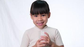 menina bonita asiática bebendo leite de um copo e sorria em fundo branco no estúdio. alimentação saudável para crianças pequenas.