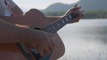 Handsome curly haired Asian man playing guitar and singing by the lake at sunset on vacation. video