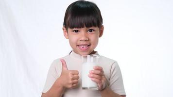 menina bonita asiática bebendo leite de um copo e mostrando o polegar para cima o sinal no fundo branco no estúdio. alimentação saudável para crianças pequenas. video