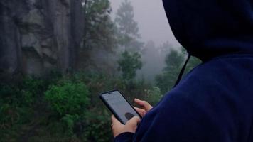 ung man resenär i marinblå manlig huvtröja lång ärm med hjälp av en smartphone och njut av landskapet i bergen med tjock dimma i bakgrunden. video