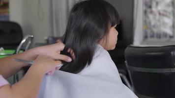 una peluquera le corta el pelo a una niña asiática en un salón de belleza. peluquero hace peinados para niñas lindas. linda niña cortando flequillo. video