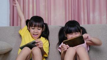 Two sisters are playing online games on their smartphones sitting on the sofa at home. Modern communication and gadget addiction concept. Two kids with gadgets. video