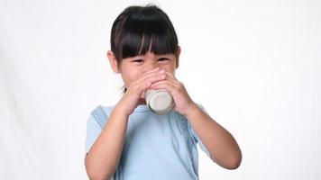 menina bonita asiática bebendo leite de um copo e sorria em fundo branco no estúdio. alimentação saudável para crianças pequenas. video