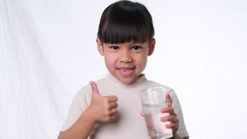 carina ragazza asiatica che beve acqua da un bicchiere e mostra i pollici in su su sfondo bianco in studio. buona sana abitudine per i bambini. concetto di assistenza sanitaria video