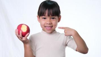 heureuse petite fille avec pomme. jolie petite fille asiatique mangeant une pomme bio sur fond blanc en studio. une alimentation saine pour les petits enfants. video