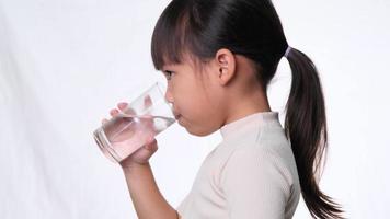 Cute little asian girl drinking water from a glass on white background in studio. Good healthy habit for children. Healthcare concept video