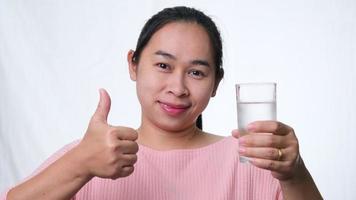 Smiling woman holding a glass of water and thumbs up on white background in studio. Healthy lifestyle video