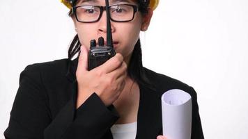 Female architect in a helmet holding construction plans and using walkie-talkie and talk to other staff on a white background in studio. video