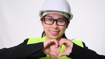 Female engineer in helmet smiling with helmet and showing heart shape with hands on white background in studio. video