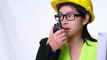 Female architect in a helmet holding construction plans and using walkie-talkie and talk to other staff on a white background in studio. video