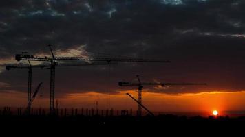 silhouette de grue à tour et chantier de construction de grands bâtiments au coucher du soleil en soirée. nouveau chantier de construction avec des grues sur fond de coucher de soleil. video