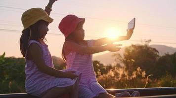 Two sisters sit on the tracks and take selfies together with their smartphones at sunset. Asian sisters enjoy spending time together on vacation. video