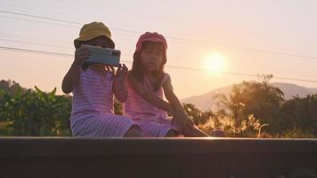 deux sœurs s'assoient sur les rails et prennent des selfies avec leurs smartphones au coucher du soleil. les sœurs asiatiques aiment passer du temps ensemble en vacances. video