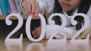 Cute little girl putting white number 2022 on a wooden table indoors. Happy new year 2022.
