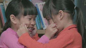 irmãs asiáticas tocam as bochechas umas das outras e sorriem cara a cara. duas meninas bonitinhas brincando juntas em casa. família adorável passando tempo juntos dentro de casa. video