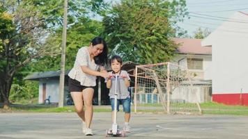 la madre le enseña a su pequeña hija a andar en scooter en el parque de verano. los niños juegan al aire libre con scooters. ocio activo y deporte al aire libre para niños video