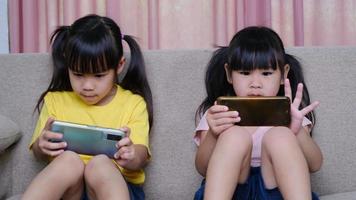 dos hermanas están jugando juegos en línea en sus teléfonos inteligentes sentadas en el sofá de casa. concepto moderno de comunicación y adicción a los gadgets. dos niños con aparatos.