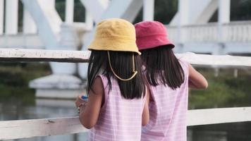 twee schattige aziatische meisjes die plezier hebben met het voeren van de vissen aan de tha-rivier. twee zussen bezoeken de oude witte spoorbrug, tha chompoo, een beroemde toeristische attractie in het noorden van thailand. video