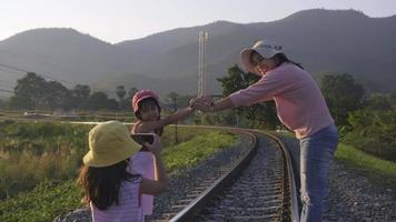 una hermana toma una foto de la madre y la hermana pequeña con un smartphone para grabar recuerdos mientras camina por las vías del tren en el campo contra las montañas por la noche. video