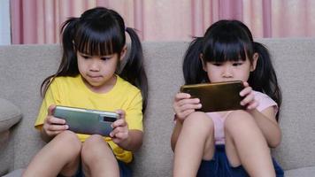 dos hermanas están jugando juegos en línea en sus teléfonos inteligentes sentadas en el sofá de casa. concepto moderno de comunicación y adicción a los gadgets. dos niños con aparatos. video