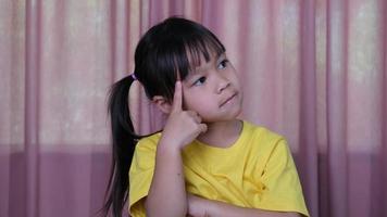 Cute little girl is thinking about something and looking up on the pink curtain background at home. video