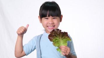 Happy little girl with fresh salad with showing thumbs up on white background in studio. Good healthy habit for children. Healthcare concept