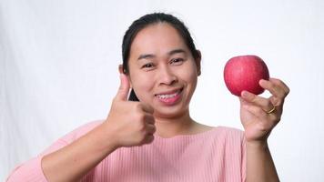 mulher asiática segurando a maçã vermelha com os polegares para cima e sorrindo mostrando dentes saudáveis em fundo branco em estúdio