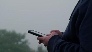 jonge man reiziger in marineblauw mannelijke hoodie sweatshirt lange mouw met behulp van een smartphone en geniet van het landschap in de bergen met dikke mist op de achtergrond. video