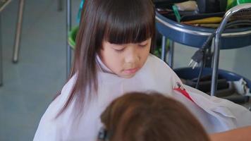 una peluquera le corta el pelo a una niña asiática en un salón de belleza. peluquero hace peinados para niñas lindas. linda niña cortando flequillo. video
